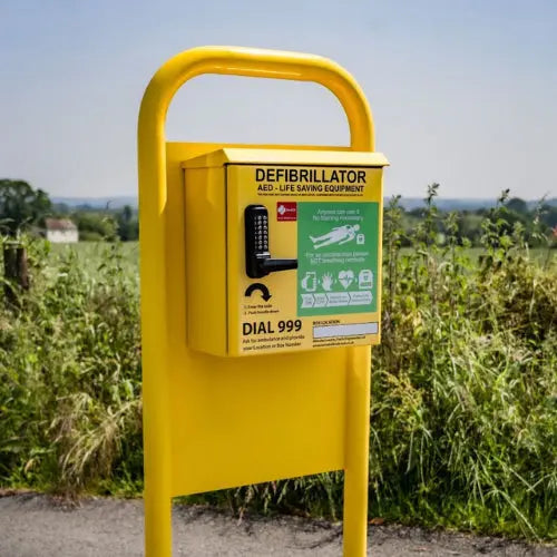 Yellow defibrillator box in a rural setting with greenery and clear sky.
